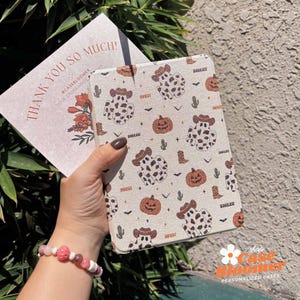 Puede incluir: Una funda beige para tableta con un patrón repetido de figuras con estampado de vaca que llevan sombreros de vaquero, calabazas y botas. La funda se sostiene con una tarjeta que dice "THANK YOU SO MUCH!" y "@CASEBLOOMER".