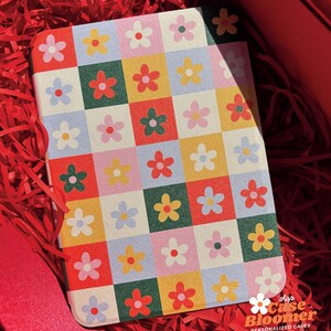 Puede incluir: Un peque&ntilde;o cuaderno con un patr&oacute;n de cuadros coloridos con flores rojas, amarillas, verdes, azules y rosas. La cubierta del cuaderno tiene el texto "Shop Case Bloomer Personalized Cases" en la parte inferior.