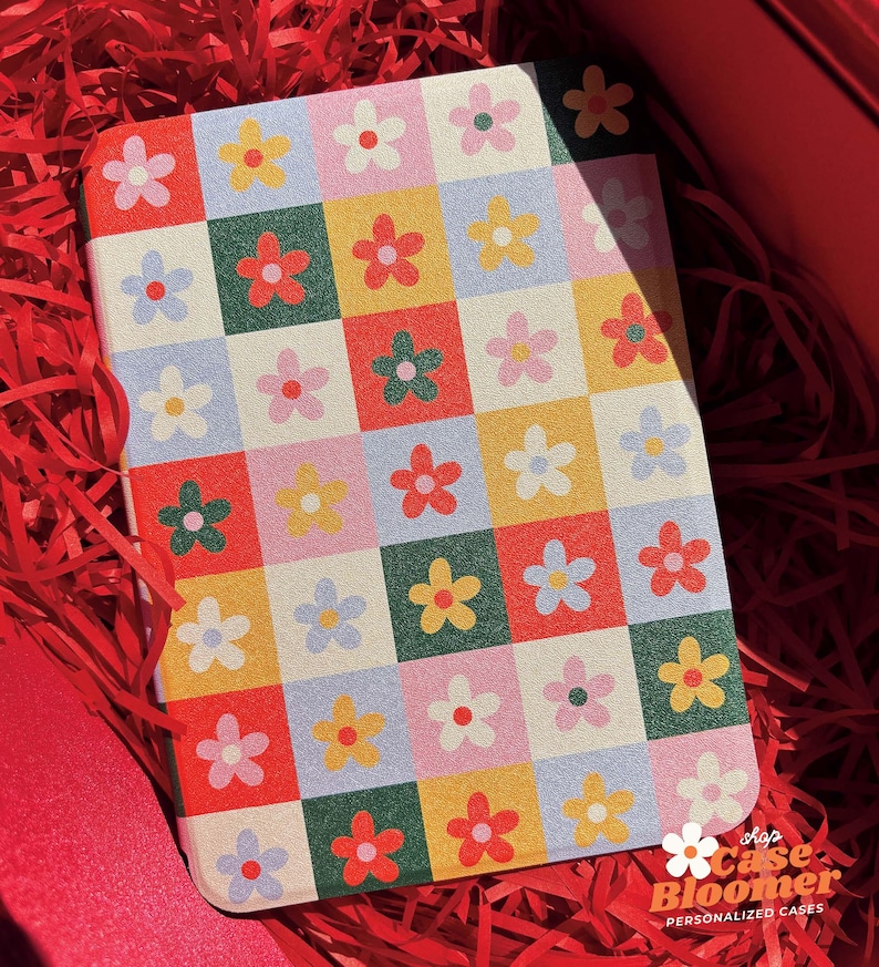 Puede incluir: Un peque&ntilde;o cuaderno con un patr&oacute;n de cuadros coloridos con flores rojas, amarillas, verdes, azules y rosas. La cubierta del cuaderno tiene el texto "Shop Case Bloomer Personalized Cases" en la parte inferior.