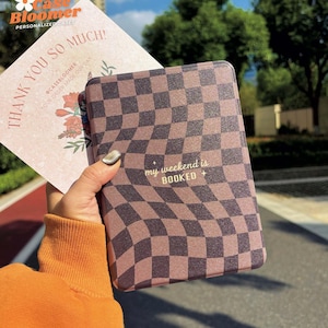 May include: A brown and black checkered tablet case with the text "my weekend is BOOKED" in white. A thank you card with floral illustrations is behind the case. The case is held in front of a blurred outdoor background.
