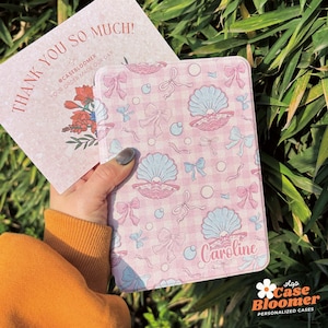 May include: A pink and white personalised phone case with a seashell and bow pattern. The case has the name "Caroline" printed on it.