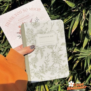 Könnte beinhalten: Ein grünes Hefter mit einem Blumendesign und dem Aufdruck "Composition Book" auf dem vorderen Umschlag. Das Heft wird in einer Hand gehalten, die einen orangebraunen Pullover trägt.