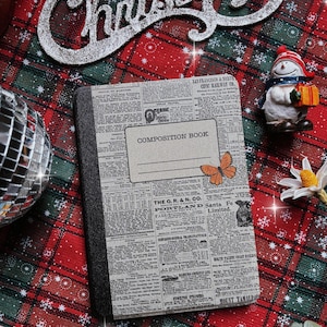 Peut inclure: Un carnet de composition avec une couverture en papier journal avec le texte "COMPOSITION BOOK" et un papillon. Le carnet est sur une surface à carreaux rouge et vert avec une inscription "Merry Christmas" en argent et une figurine de bonhomme de neige.