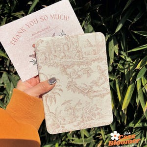 May include: A hand holds a light beige notebook with a pink floral pattern. The notebook is closed and has a white border. The background is a green leafy plant.