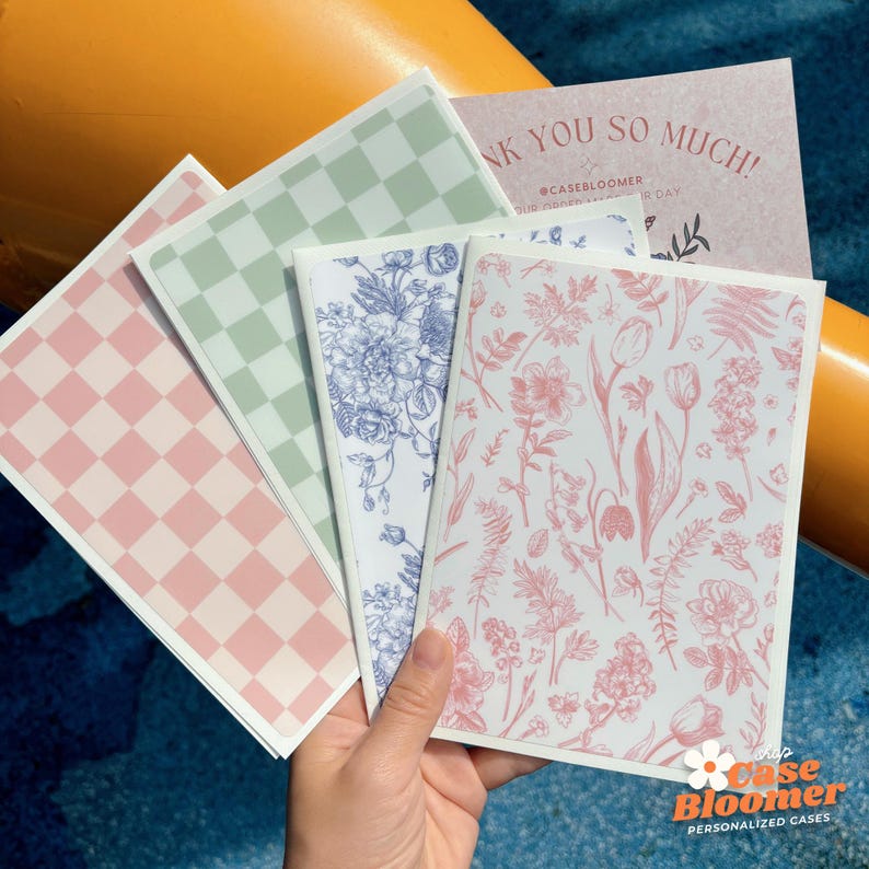 May include: A hand holds a stack of notebooks with various floral and checkered patterns. The notebooks feature pink, blue, and green designs. A thank you card is visible in the background. The brand logo is at the bottom right.