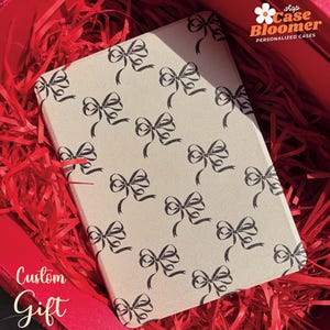 May include: A beige custom gift box with a black bow pattern. The box is filled with red shredded paper. The box has the text "Custom Gift" on the front. The box is labeled "shop Case Bloomer PERSONALIZED CASES".