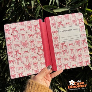 May include: A pink and white patterned Kindle cover with a black spine. The cover has a repeating pattern of pink bows on a white background. The spine has the text "COMPOSITION BOOK" printed on it.