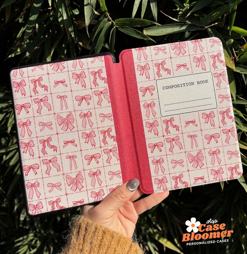 May include: A pink and white patterned Kindle cover with a black spine. The cover has a repeating pattern of pink bows on a white background. The spine has the text "COMPOSITION BOOK" printed on it.