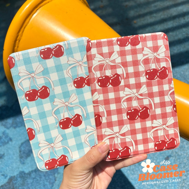 Puede incluir: Dos fundas para tel&eacute;fono con un patr&oacute;n de cuadros vichy y cerezas. La funda de la izquierda es azul y blanca, y la funda de la derecha es roja y blanca.