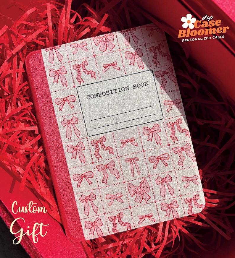 May include: A red composition book with a white cover featuring a repeating pattern of pink bows. The cover also has the text "COMPOSITION BOOK" printed in black.