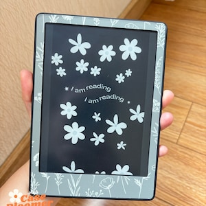 May include: A gray e-reader case with a floral design and the text "I am reading." The case features white flowers and botanical illustrations. The e-reader is held in a hand, showcasing the design and the screen.
