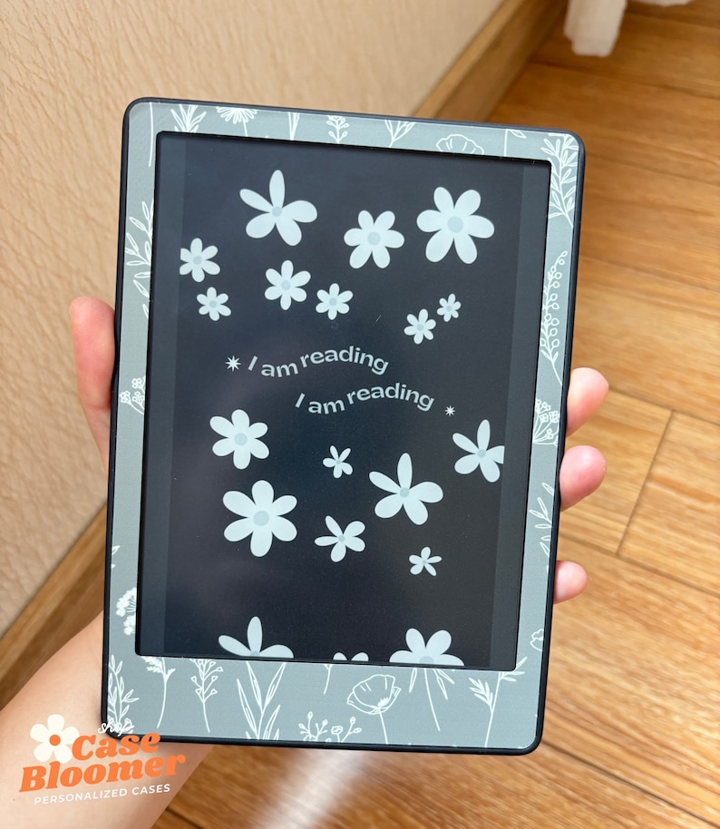 May include: A gray e-reader case with a floral design and the text "I am reading." The case features white flowers and botanical illustrations. The e-reader is held in a hand, showcasing the design and the screen.