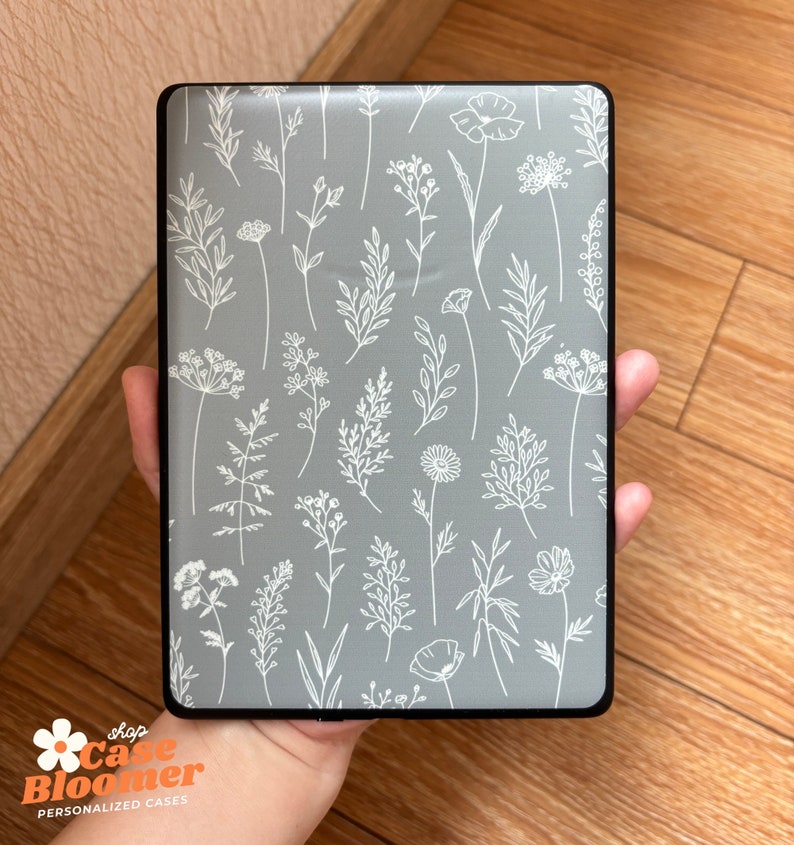 May include: A rectangular case with a gray background and a white floral pattern. The case has a black border and is held in a hand. The text "shop Case Bloomer PERSONALIZED CASES" is visible in the lower left corner.