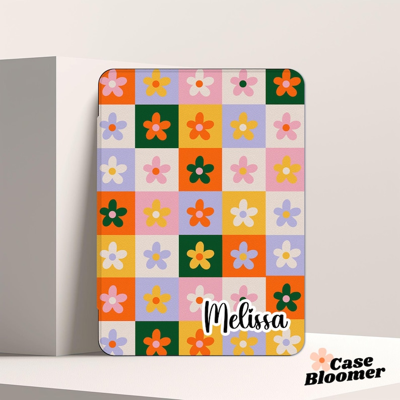 Puede incluir: Una funda de tel&eacute;fono personalizada con un patr&oacute;n de cuadros coloridos con flores en tonos naranja, amarillo, rosa y verde. El nombre "Melissa" est&aacute; impreso en negro en la funda.