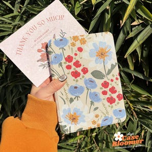 May include: A floral patterned notebook cover with blue, red and yellow flowers on a cream background. The text "Shop Case Bloomer Personalised Cases" is at the bottom of the cover.