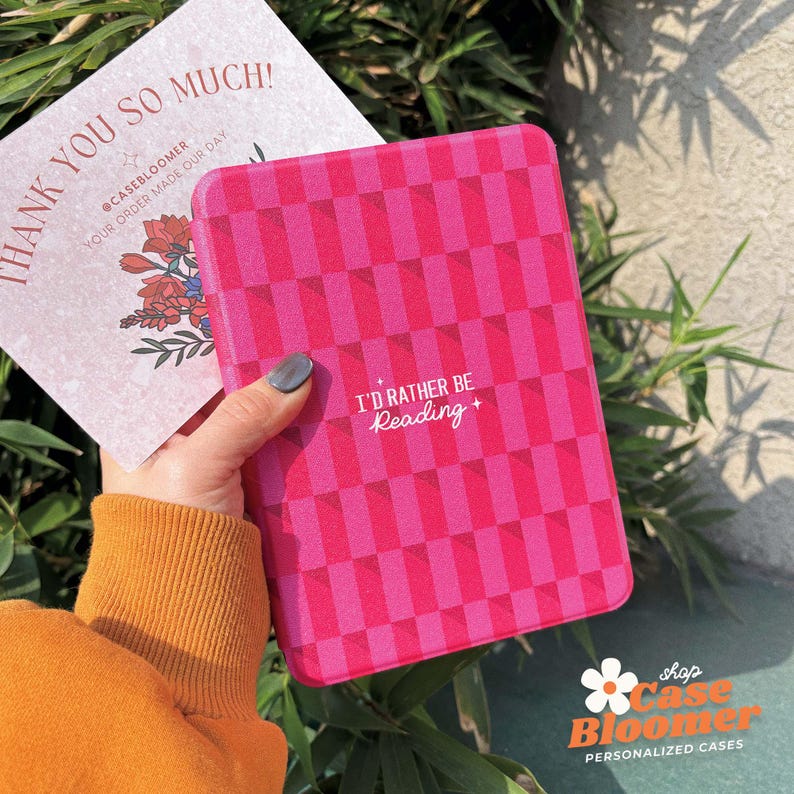 May include: A pink and white chequered e-reader case with the text "I'd rather be reading +" in white. The case is made of a soft, durable material and is designed to protect your e-reader from scratches and bumps.