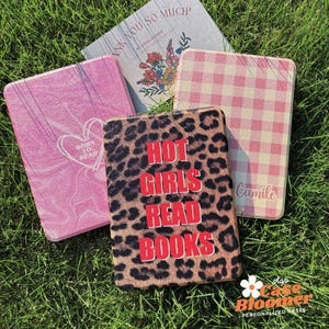May include: Several personalised cases are displayed, each with a unique design. One case has a leopard print with the text "HOT GIRLS READ BOOKS" in red. Other cases feature pink marble, a pink gingham pattern, and a floral design with the text "THANK YOU SO MUCH!"