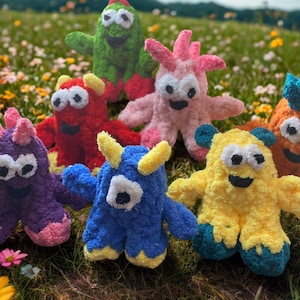 May include: A collection of handmade plush monster toys in various colors, including purple, red, green, pink, blue, orange, and yellow. Each monster has a unique design with different horn and eye shapes, set against a backdrop of wildflowers.
