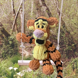 May include: A plush, orange and black tiger toy with a yellow belly and a purple nose sits on a small wooden swing. The toy is suspended by white rope, set against a blurred green forest background.