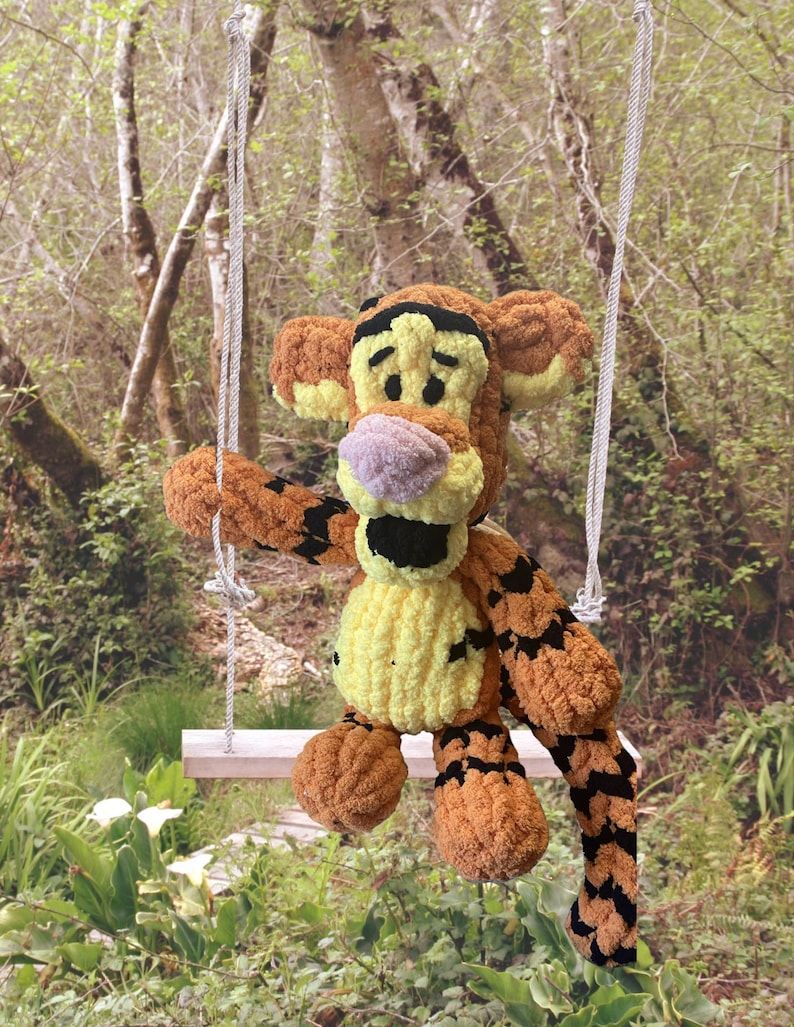 May include: A plush, orange and black tiger toy with a yellow belly and a purple nose sits on a small wooden swing. The toy is suspended by white rope, set against a blurred green forest background.