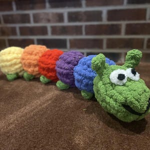 May include: A colorful, crocheted caterpillar toy in a rainbow of colors: yellow, orange, red, purple, blue, and green. The caterpillar has large white eyes with black pupils and a smiling mouth. The toy is made of soft yarn.