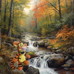 May include: A vibrant, hand-crocheted Tigger plush toy stands on a rock in front of a scenic painting of a river and waterfall. The plush features orange and yellow yarn with black stripes, a pink nose, and a black mouth. The background depicts an autumn forest.