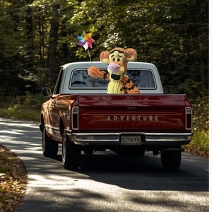 May include: A red vintage pickup truck with a white cab drives down a tree-lined road. A plush Tigger doll holds a colorful pinwheel in the truck bed. The word "ADVENTURE" is printed across the tailgate.