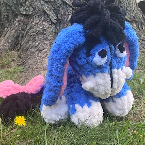 May include: A plush blue donkey toy with white accents on its face and feet, black hair, and pink inner ears. A pink and black bow is next to the donkey on the grass. A yellow dandelion is also visible.