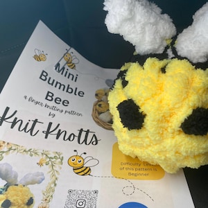 May include: A handmade yellow and black bumble bee toy with white wings rests on a printed pattern sheet. The sheet features the text "Mini Bumble Bee" and "Knit Knotts", along with a bee illustration and a QR code.