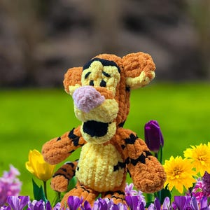 Può includere: Un peluche di Tigger all'uncinetto arancione e giallo fatto a mano con strisce nere, in piedi in un campo di fiori viola, gialli e rosa. Il peluche ha un naso rosa e dettagli neri. Lo sfondo è verde sfocato.