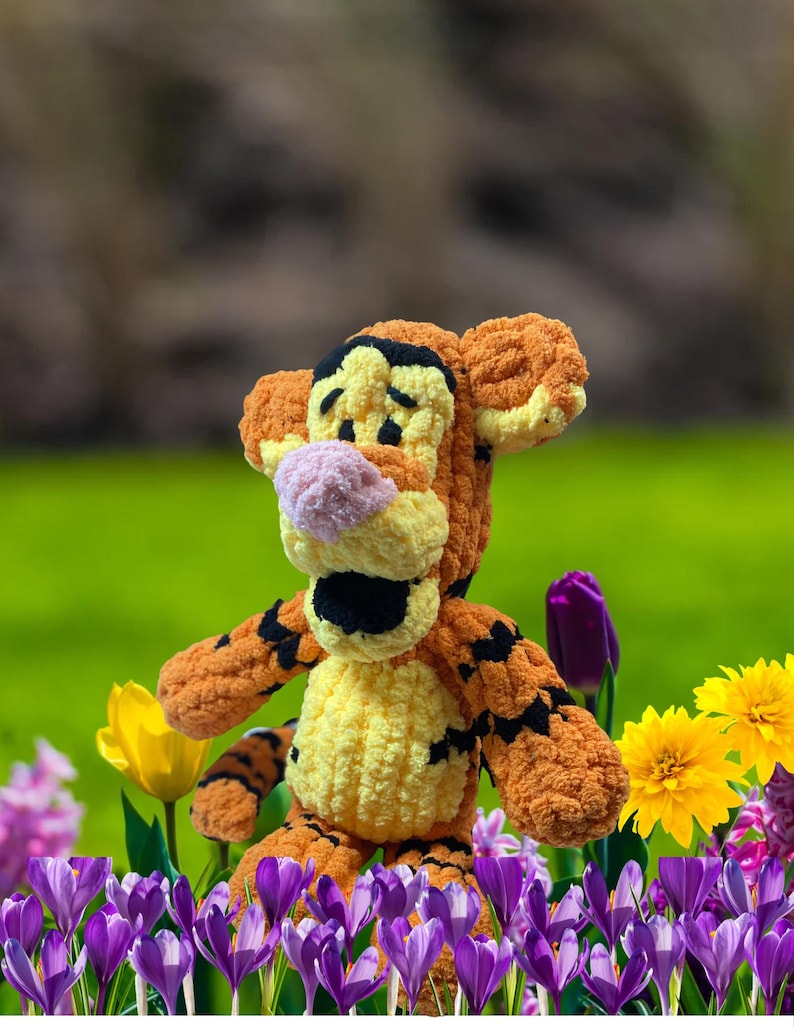 May include: A handmade orange and yellow crochet Tigger plush toy with black stripes, standing in a field of purple, yellow, and pink flowers. The plush has a pink nose and black accents. The background is a blurred green.