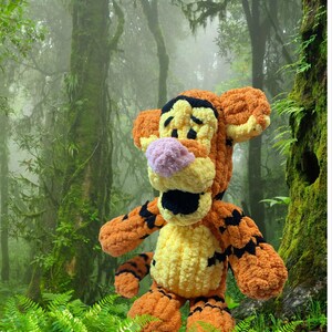 May include: A handmade, plush Tigger toy, featuring orange, yellow, black, and pink colors. The stuffed animal has a smiling expression and is set against a lush, green forest background. The toy is a character from the Winnie the Pooh series.
