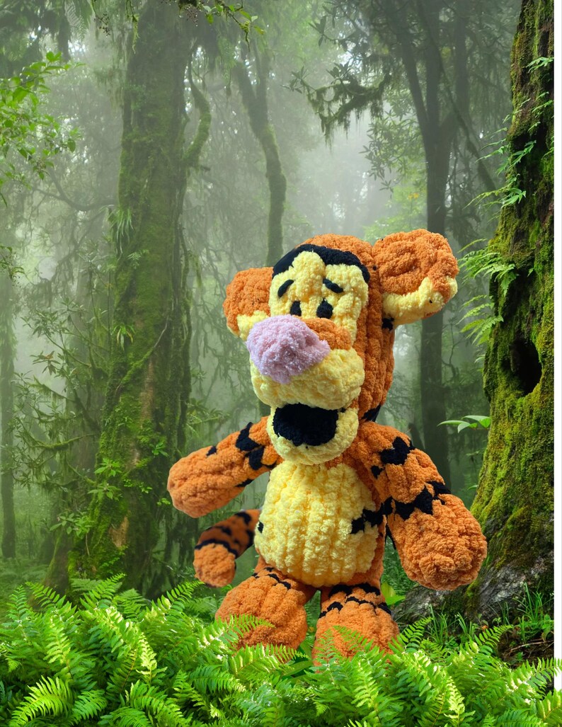 May include: A handmade, plush Tigger toy, featuring orange, yellow, black, and pink colors. The stuffed animal has a smiling expression and is set against a lush, green forest background. The toy is a character from the Winnie the Pooh series.