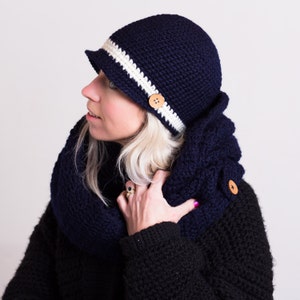 May include: A navy blue crocheted hat with a white stripe and a wooden button. The hat has a wide brim and is worn by a person with blonde hair.