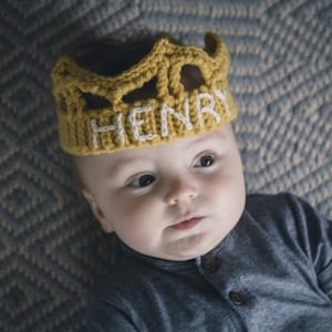 May include: A baby wearing a yellow crocheted crown with the name "HENRY" embroidered in white.