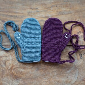 Two crocheted mittens with smiling faces. One is grey and the other is purple, plum.