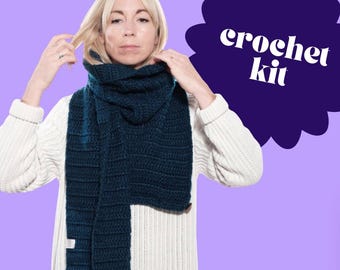 Puede incluir: Se muestra una bufanda de ganchillo de color verde azulado oscuro. La bufanda es gruesa y parece hecha a mano. Las palabras "crochet kit" están en texto blanco sobre un fondo púrpura. La persona de la imagen lleva un jersey blanco.