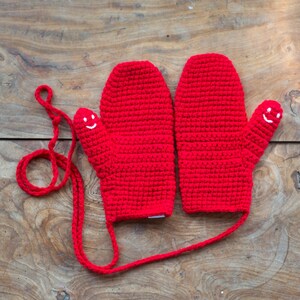 May include: A pair of red crocheted mittens with a smiling face on each thumb. The mittens are connected by a red string.