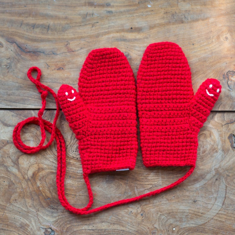 May include: A pair of red crocheted mittens with a smiling face on each thumb. The mittens are connected by a red string.