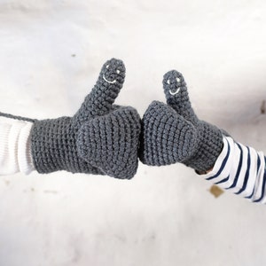 Two grey gray crocheted mittens with smiling faces. The mittens are made with a chunky yarn and have a textured surface. Mummy and me adult and child mittens showing the smiley faces embroidered onto the thumbs of the mittens.
