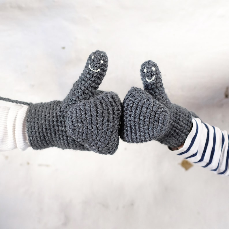 Two grey gray crocheted mittens with smiling faces. The mittens are made with a chunky yarn and have a textured surface. Mummy and me adult and child mittens showing the smiley faces embroidered onto the thumbs of the mittens.