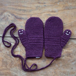 May include: A pair of purple crocheted mittens with a white smiley face on each thumb. The mittens are connected by a purple string.