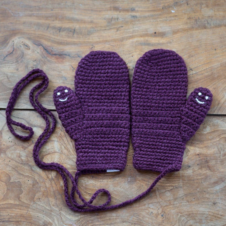 May include: A pair of purple crocheted mittens with a white smiley face on each thumb. The mittens are connected by a purple string.