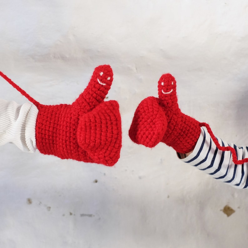 Two red crocheted mittens with smiling faces. The mittens are made with a chunky yarn and have a textured surface. Mummy and me adult and child mittens showing the smiley faces embroidered onto the thumbs of the mittens.