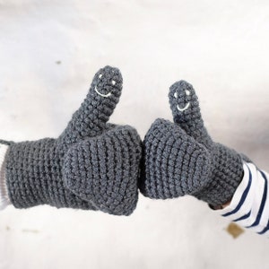 May include: A pair of gray crocheted mittens with smiley faces on the thumbs. The mittens are shown with the thumbs up.