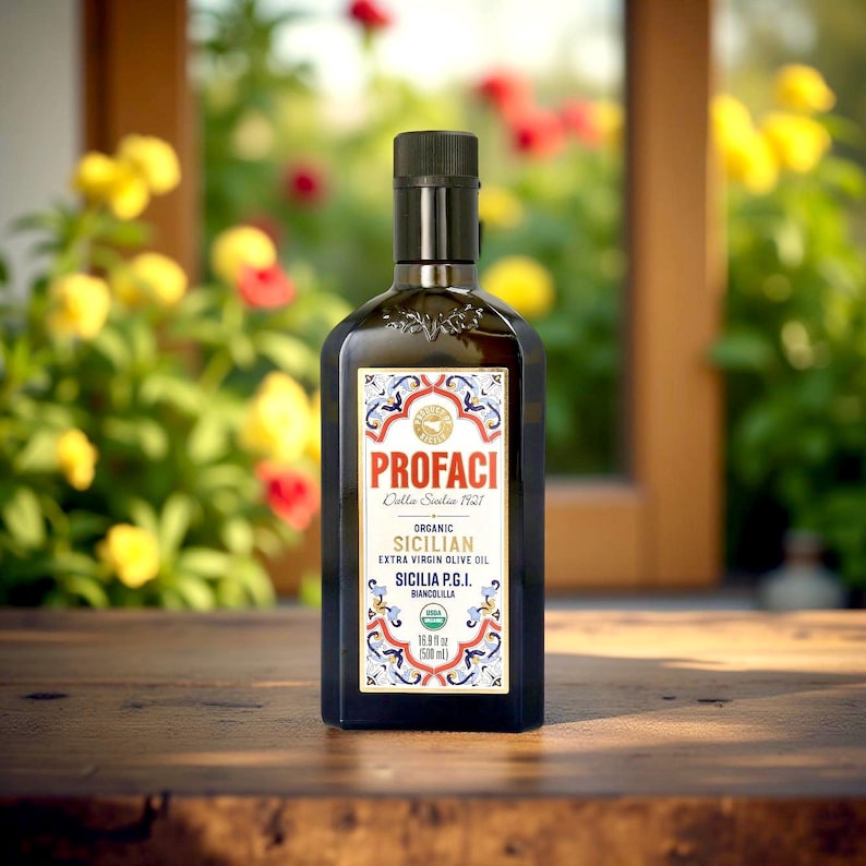May include: A glass bottle of organic Sicilian extra virgin olive oil with a gold label featuring the brand name "Profac", the text "Dalla Sicilia 1921", and the words "Sicilia P.G.I. Biancolilla". The bottle is sitting on a wooden surface.