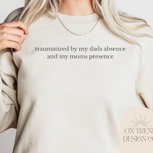 Puede incluir: Sudadera color crema con el texto negro "traumatized by my dads absence and my moms presence". La sudadera tiene cuello redondo y mangas largas. El logo de la marca "ON TREND DESIGN CO." está en la parte inferior derecha.