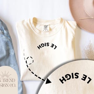 May include: A white t-shirt with a black graphic that says "Le Sigh" in a mirrored font. The graphic is surrounded by a dashed line and an arrow pointing to the right.