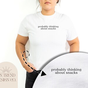 Pode incluir: Camiseta branca com o texto "probably thinking about snacks" impresso na frente.