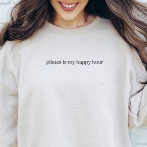 May include: A cream-coloured sweatshirt featuring the text "pilates is my happy hour" in black. The sweatshirt has a crew neck and long sleeves. The image shows the top half of a person wearing the sweatshirt.